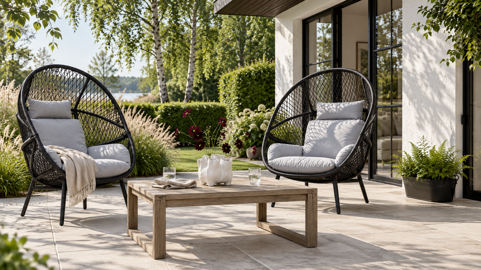 Helle Terrasse im nordischen Stil mit schwarzen Rosario Loungesesseln, Holztisch, weißer Vase und Gräsern